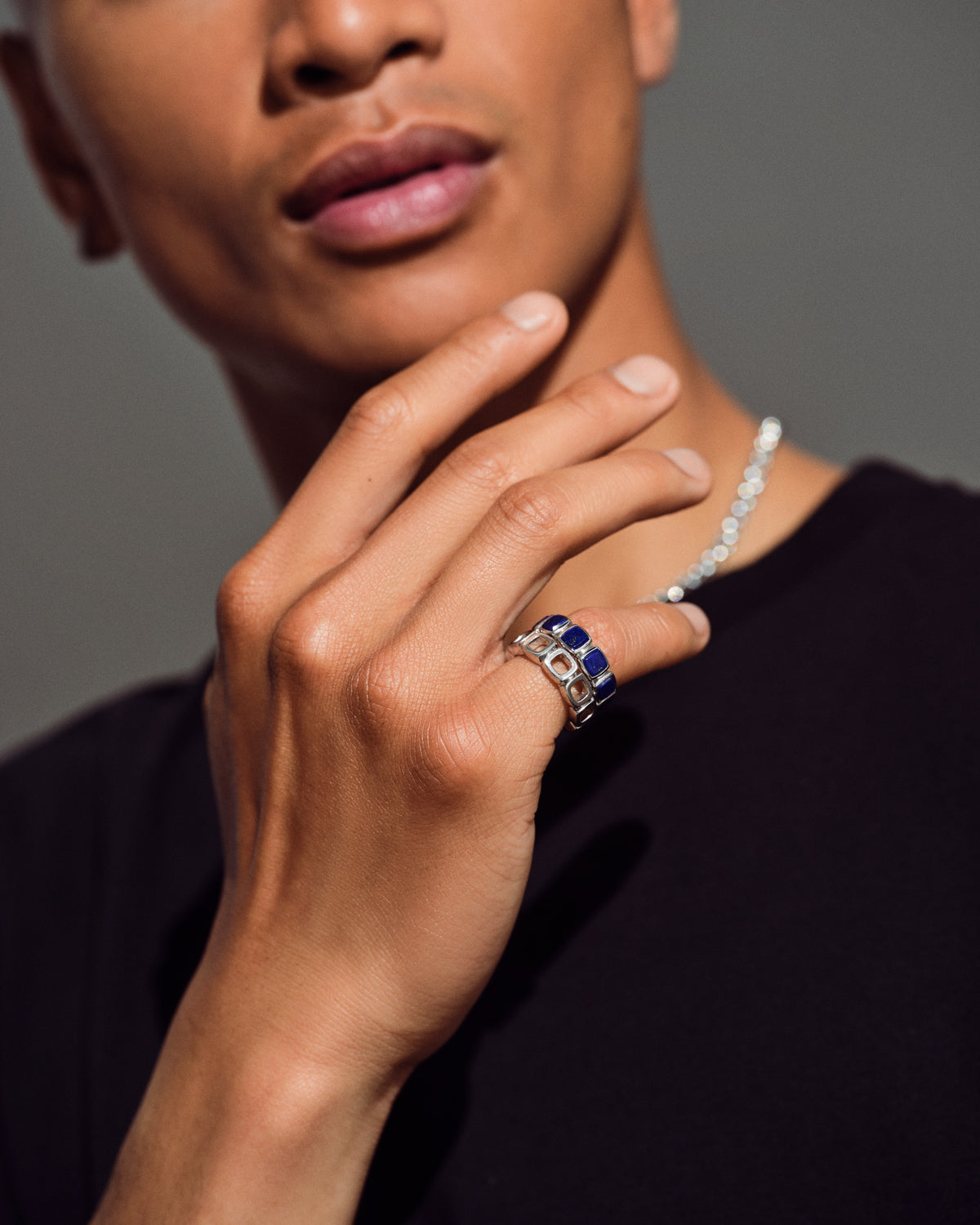 A close-up lifestyle shot of a model wearing a silver ring with blue and white stones on their ring finger, along with a silver beaded necklace.