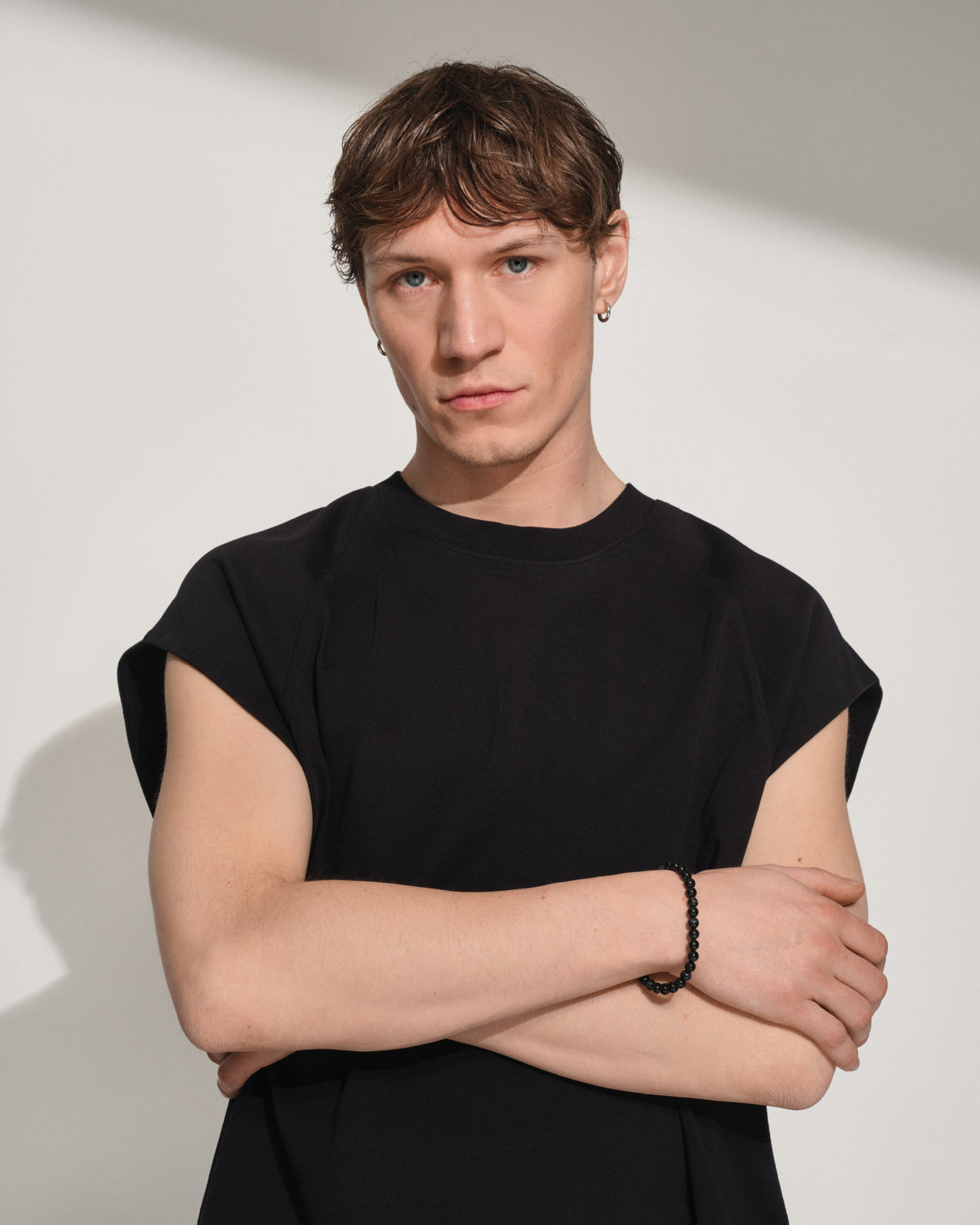 A young man with short brown hair wearing a black sleeveless t-shirt and a black beaded bracelet, posing with his arms crossed against a minimalist off-white background with soft diagonal shadows.
