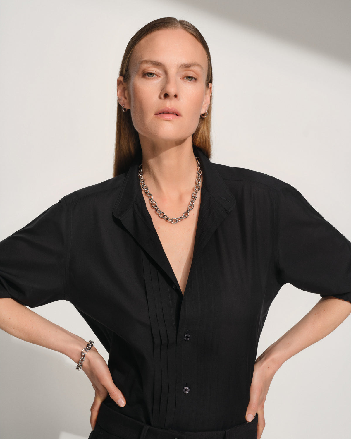 A woman modeling a black short-sleeve button-front shirt with a relaxed collar, paired with a chunky silver chain necklace and bracelet, against a light gray studio background.