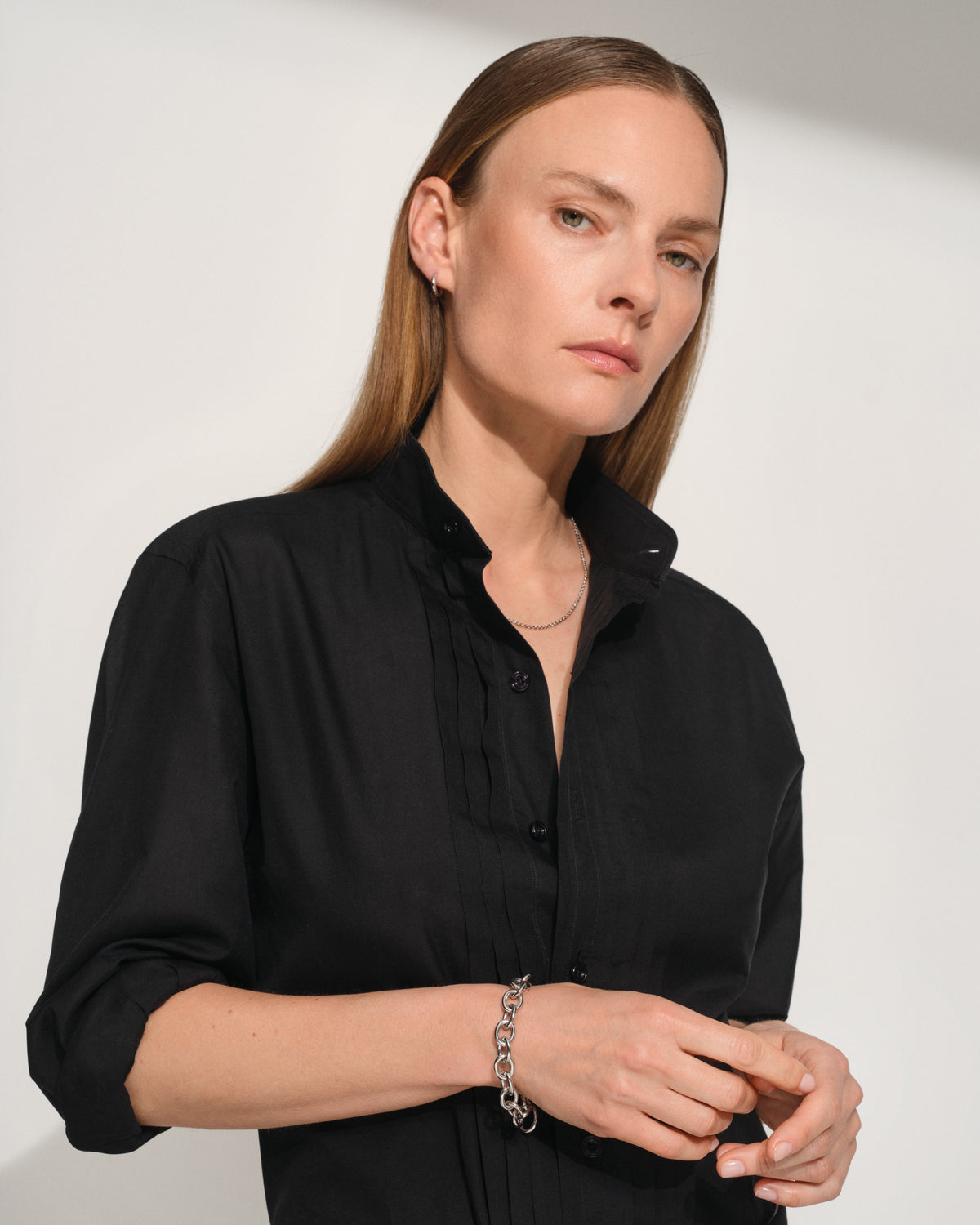 A woman wearing a black button-up shirt with pleated detailing on the chest and sleeves, with A polished silver chain link bracelet with a flat, wide oval link design, displayed on a light gray background.