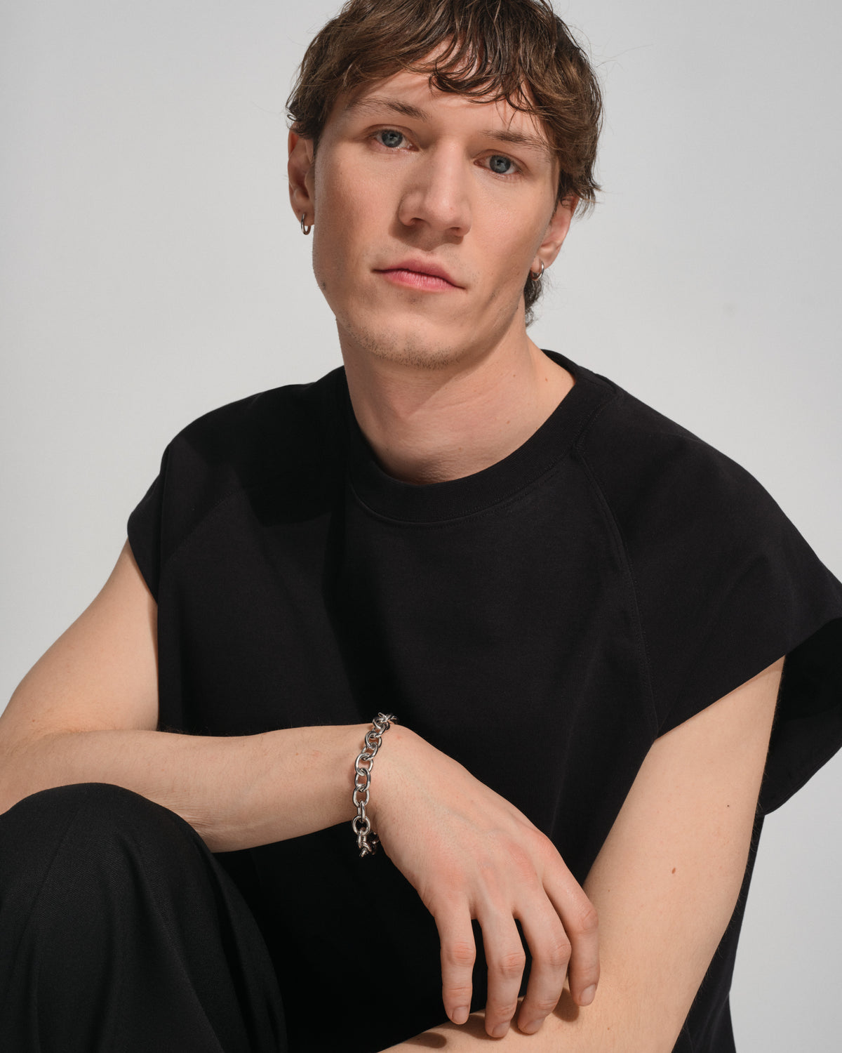 A young man with short brown hair wearing a black sleeveless t-shirt and black pants, sitting against a light gray background. He is wearing a silver chain bracelet on his wrist.