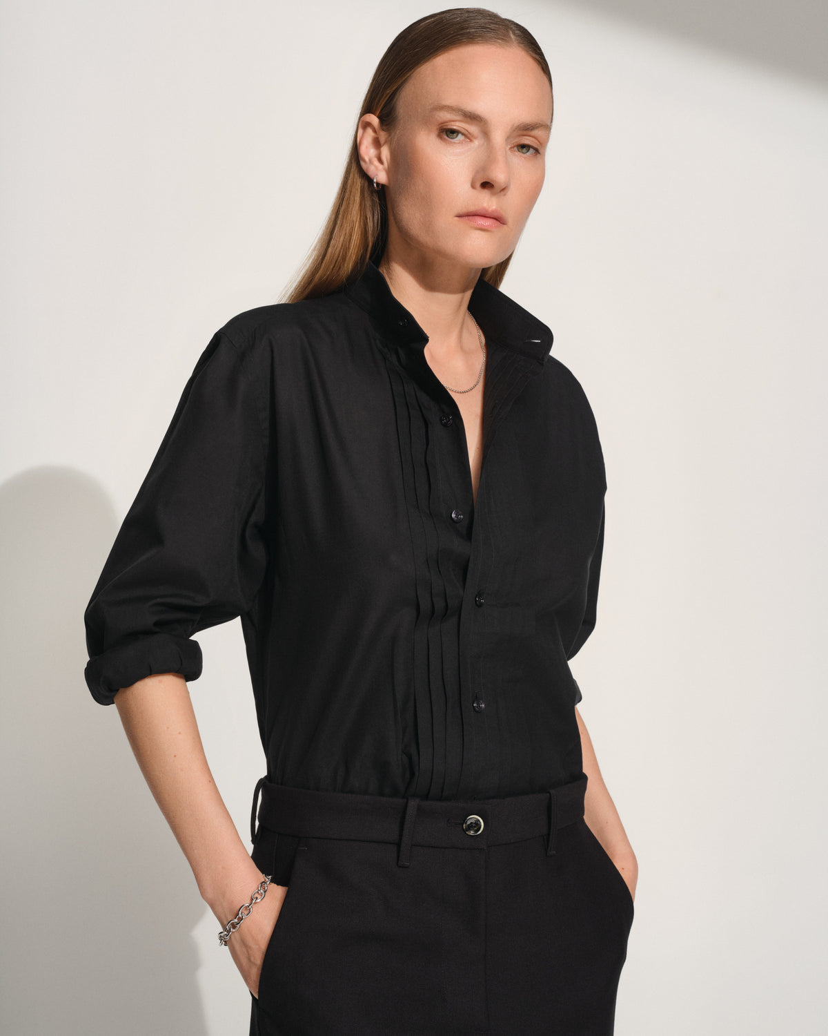 A woman modeling a black long-sleeve button-up shirt with pleated detailing on the front, paired with black trousers, silver bracelet and necklace against a light gray studio background.