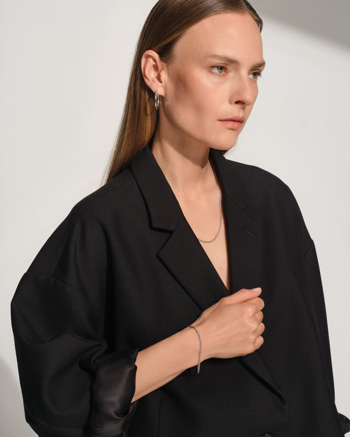 A woman wearing a black oversized blazer with wide sleeves, styled with delicate silver jewelry including a thin chain necklace, bracelet, and hoop earrings, set against a minimalist light gray background.