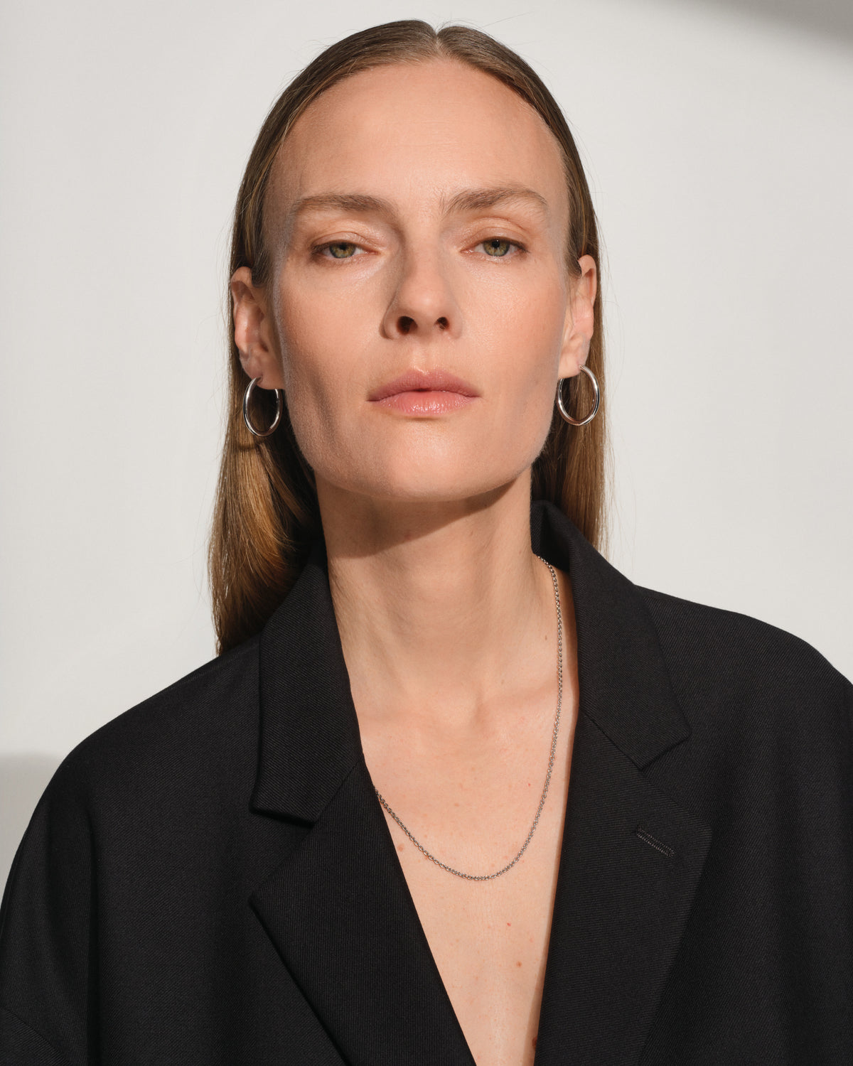 A close-up portrait of a woman with straight light brown hair wearing silver hoop earrings, a delicate silver necklace, and a black blazer against a plain light gray background.