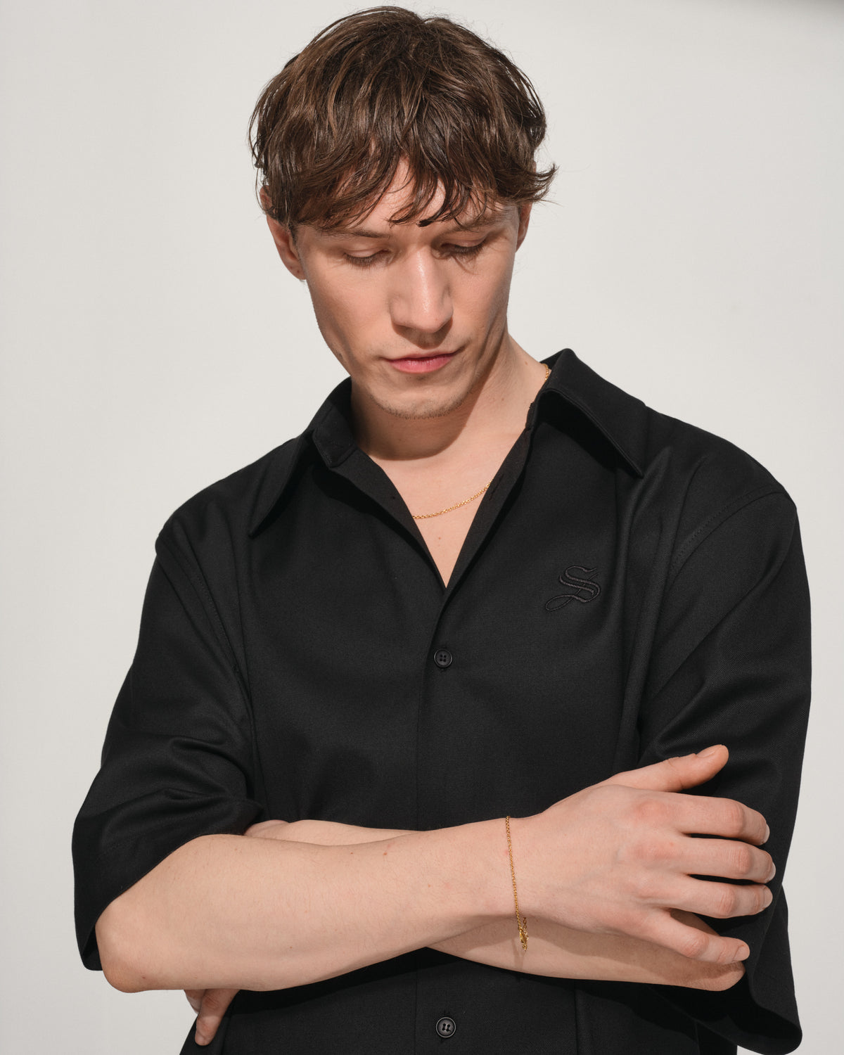 A man wearing a black button-up shirt with a small ada super slim gold necklace, posed with arms crossed against a light gray background.