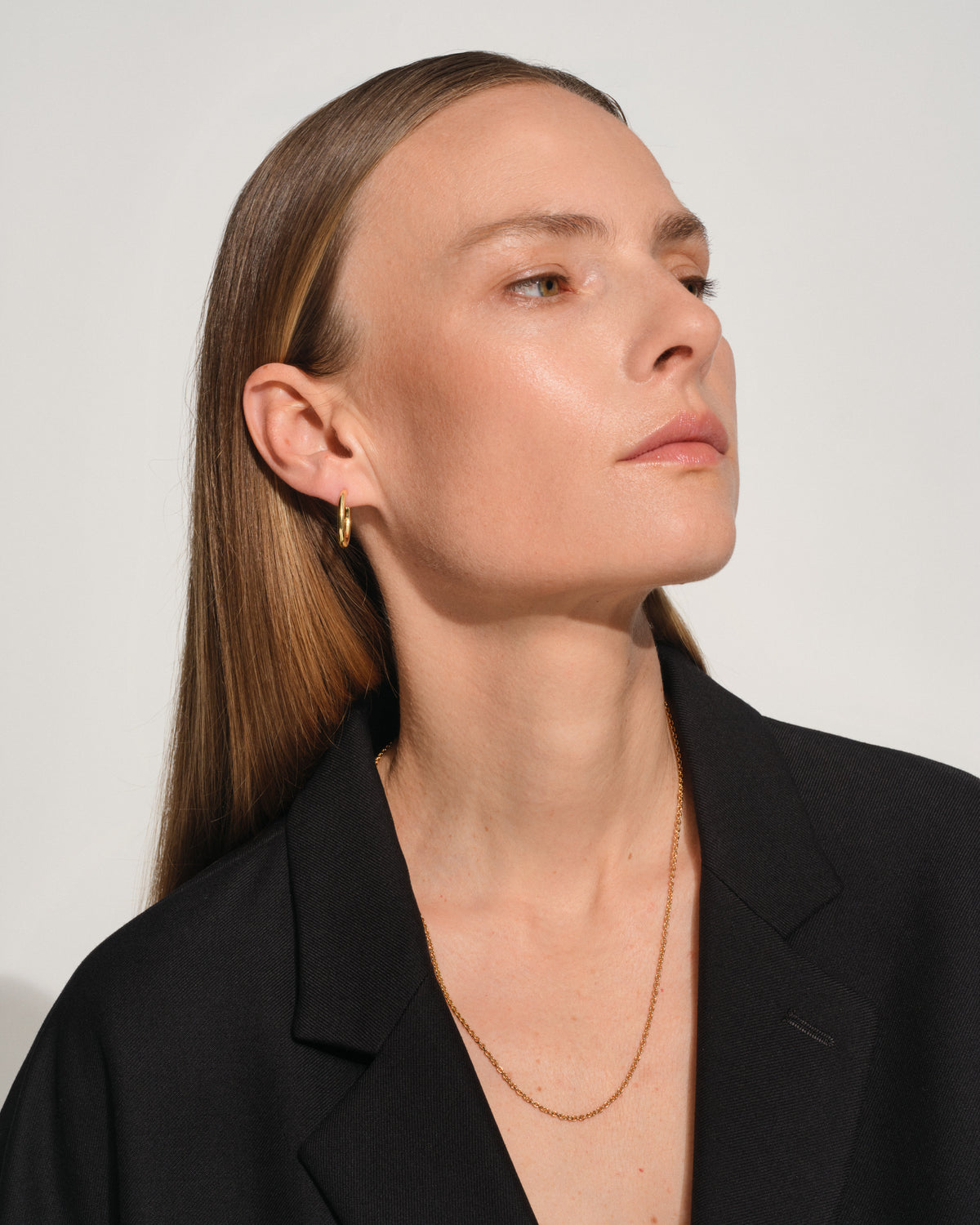A close-up portrait of a woman wearing a delicate gold chain necklace and a small gold hoop earring, dressed in a black blazer against a plain light gray background.