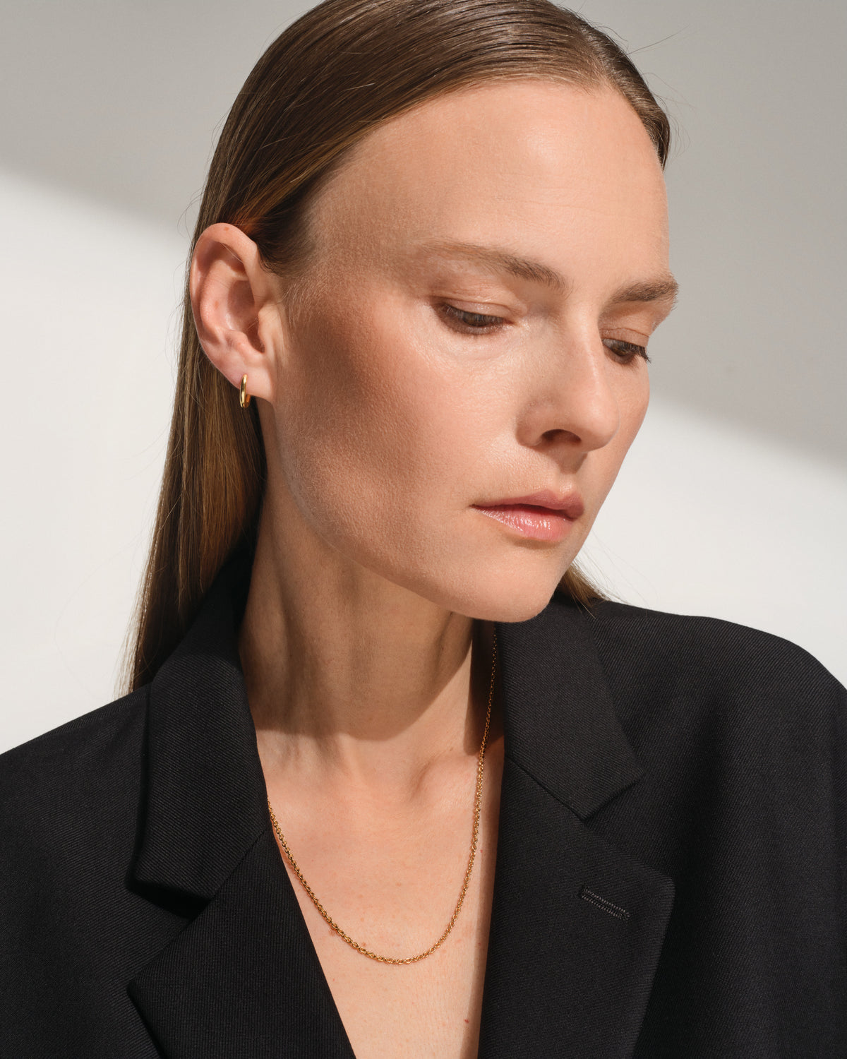 A close-up of a woman wearing a delicate ada chain super slim gold chain necklace and a small gold hoop earring, dressed in a black blazer against a neutral light gray background.