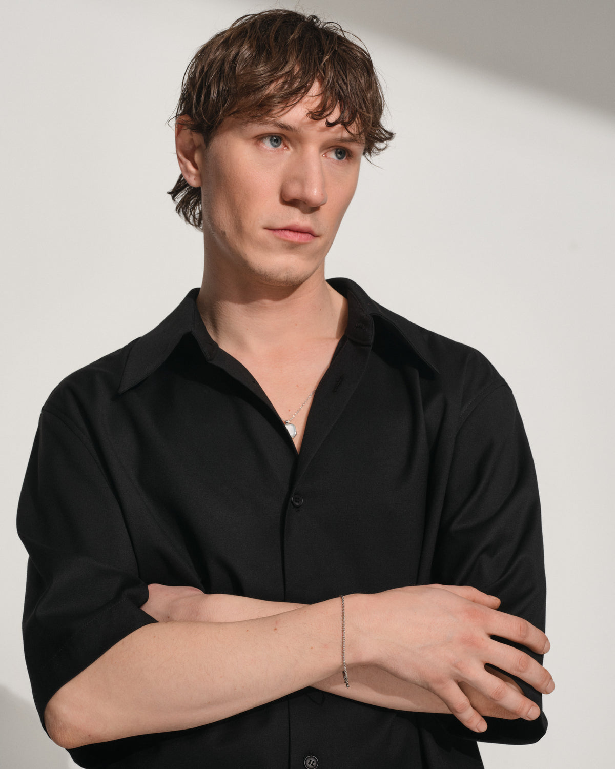 A man with light brown hair wearing a black button-down shirt, standing with his arms crossed against a plain light gray background.