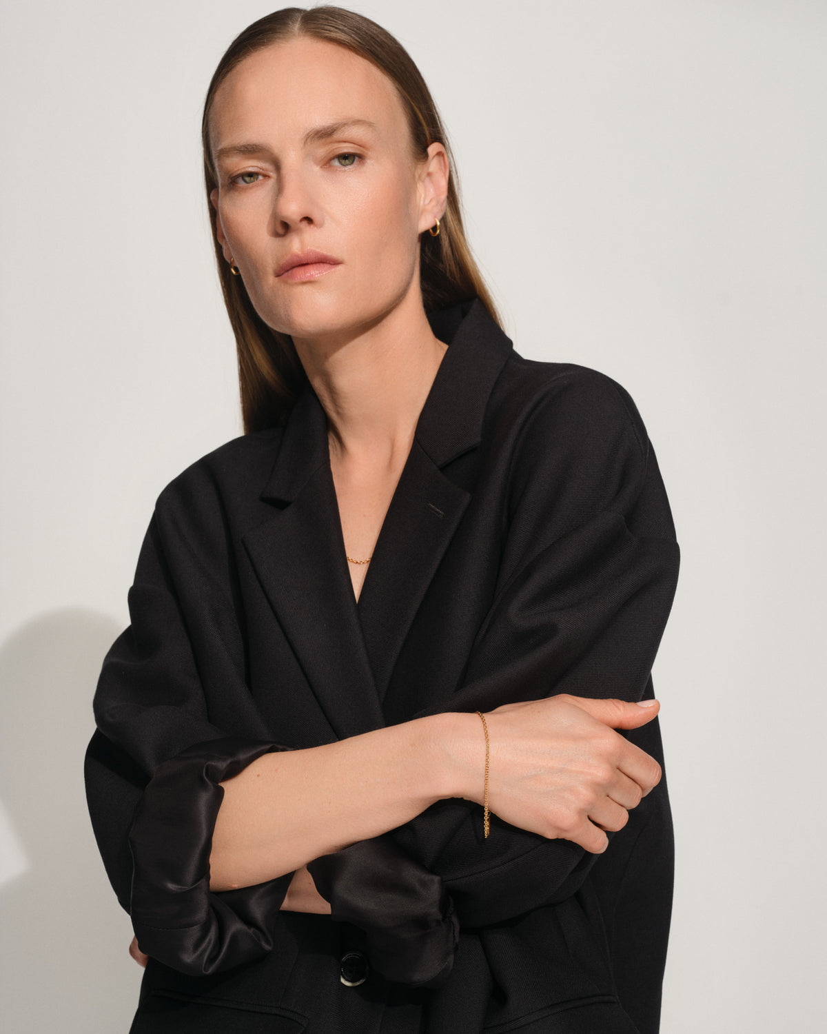 A woman with long light brown hair wearing a black satin-trimmed blazer and gold jewelry, posing with her arms crossed against a light gray studio background.