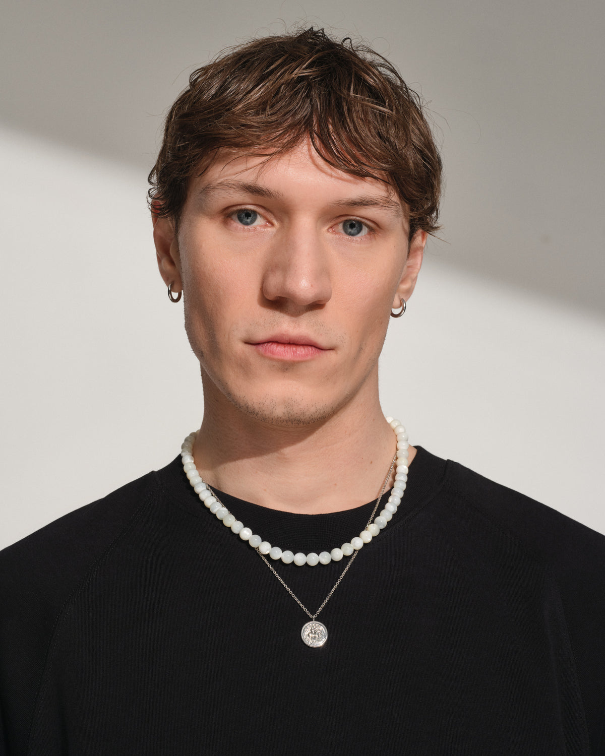 Tom Wood Cove Necklace White MOP - A man wearing layered necklaces. One necklace is a classic white pearl strand, and the other is a silver chain with a round silver pendant.