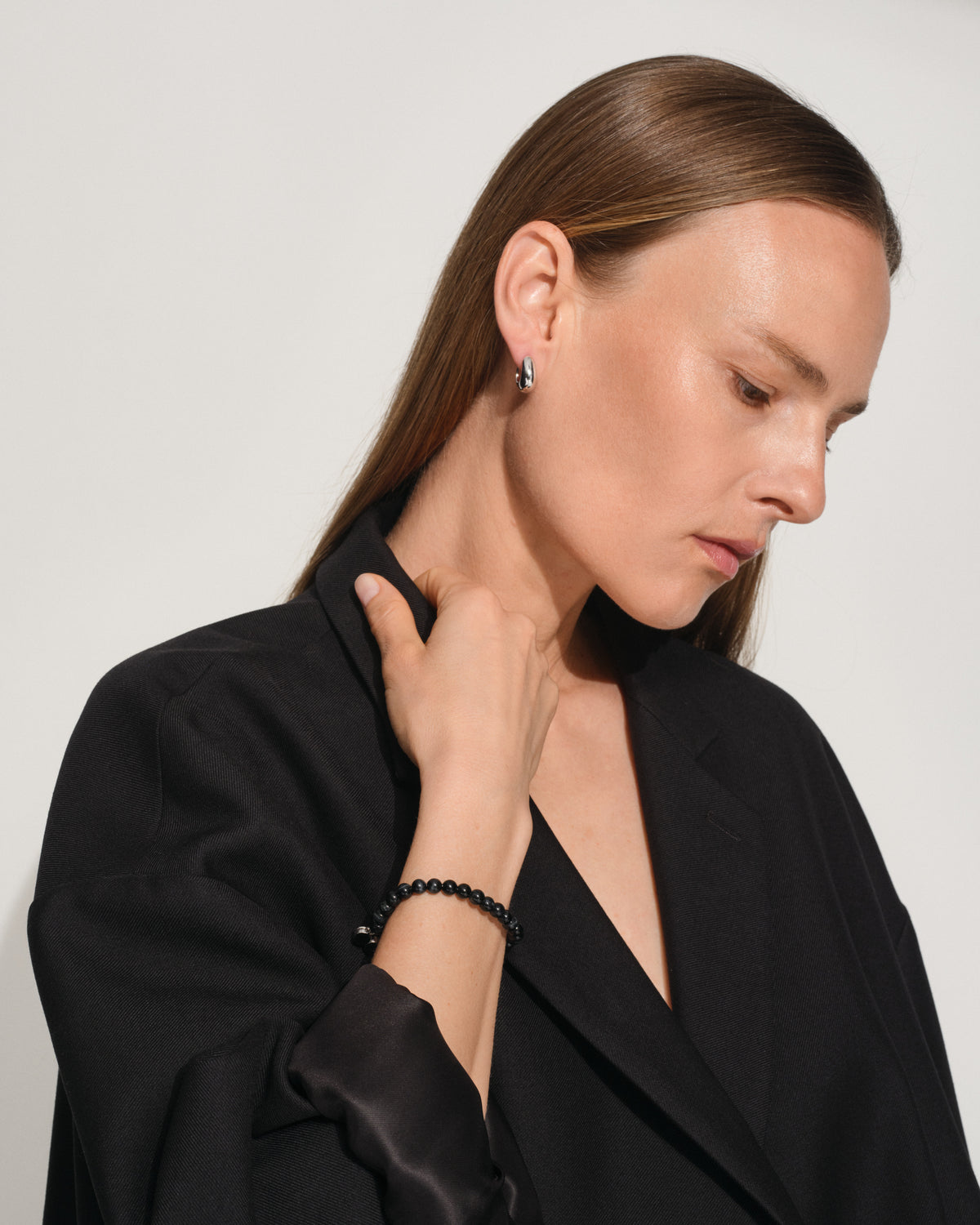A woman with long brown hair, wearing a black satin blazer, is shown in profile looking down. She is wearing a black beaded bracelet on her wrist and a small silver hoop earring.