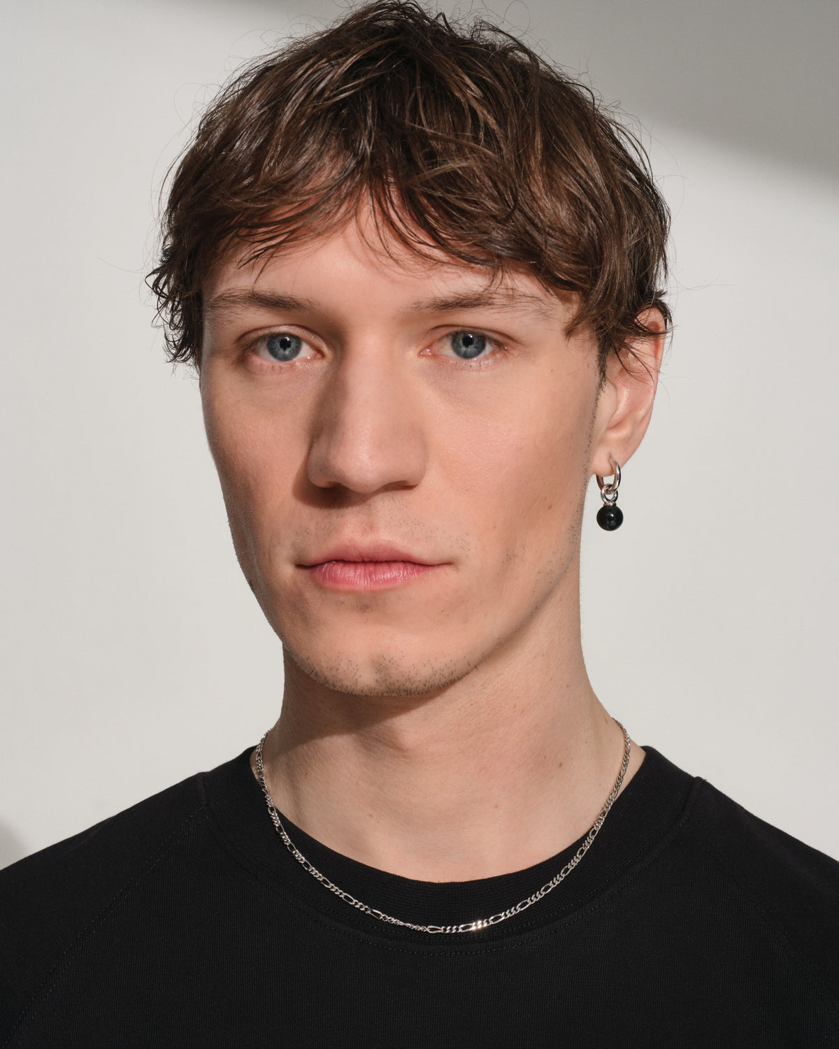 A close-up portrait of a man with light blue eyes, brown hair, and a black crewneck shirt, wearing a silver chain necklace and a black dangle earring against a light gray background.