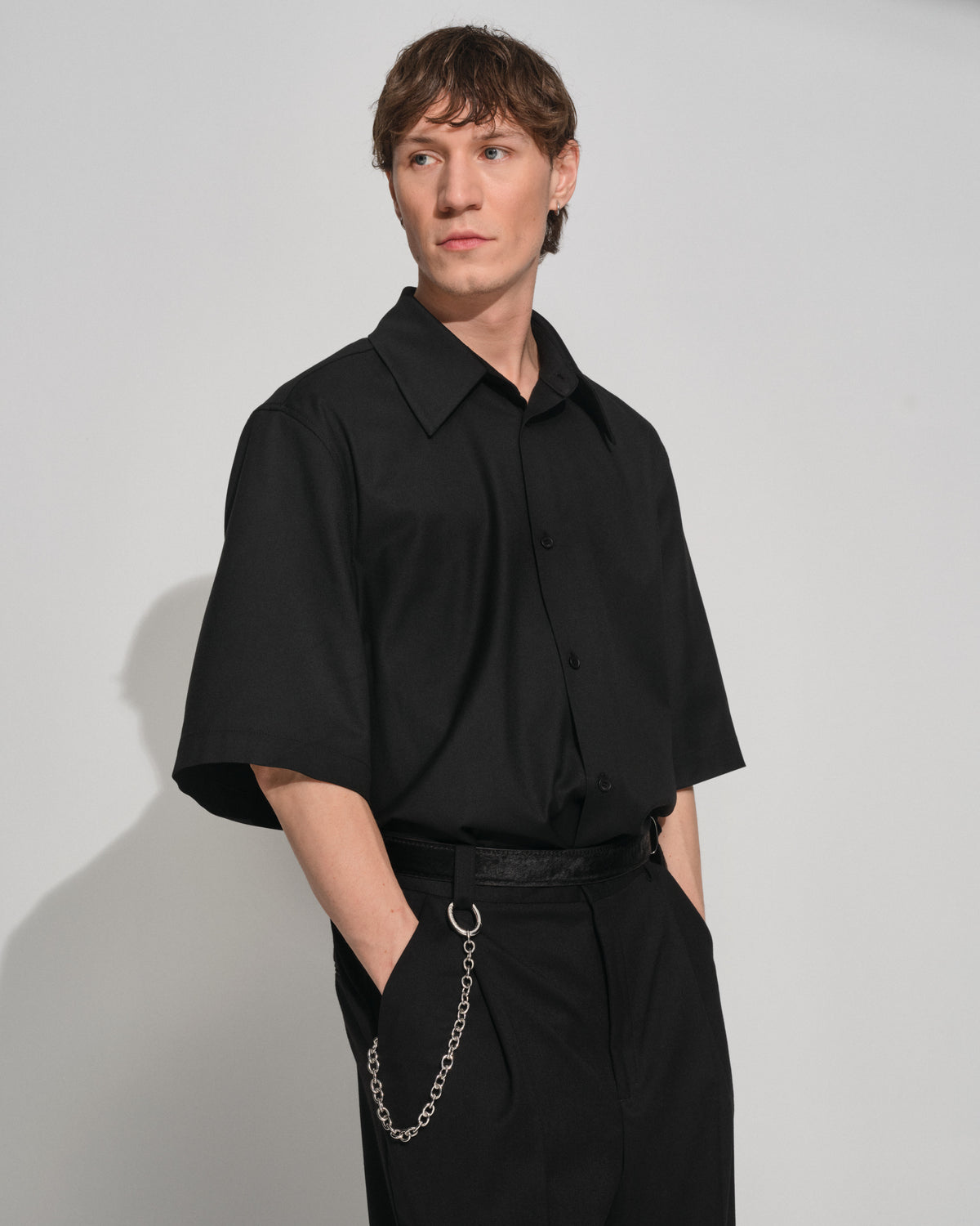 A man wearing a black short-sleeve button-up shirt and black trousers, styling a silver key clasp standing against a light gray studio background with one hand in his pocket.