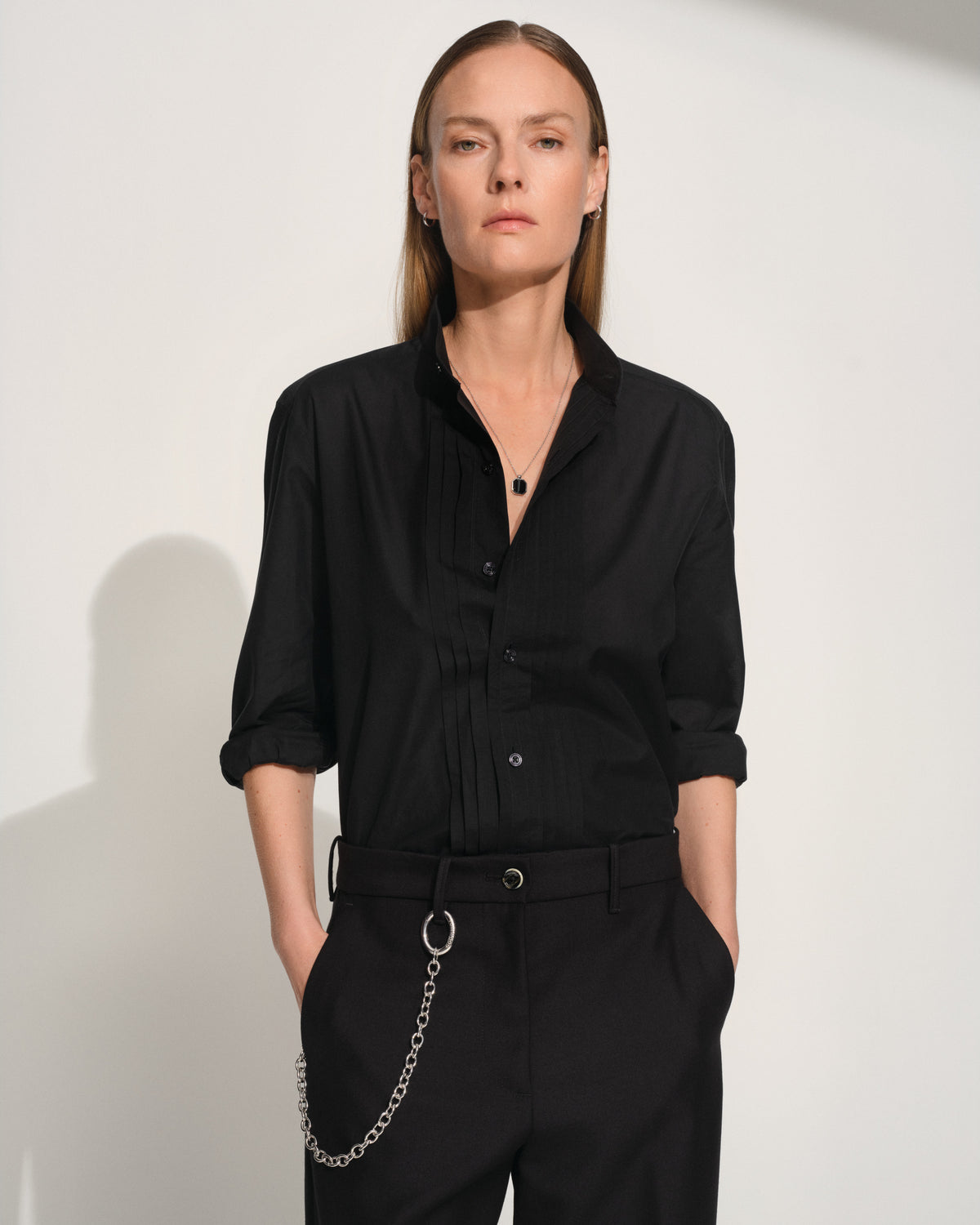 A woman modeling a black long-sleeve button-up shirt with rolled sleeves and black trousers styling a silver key clasp, standing against a plain light background.