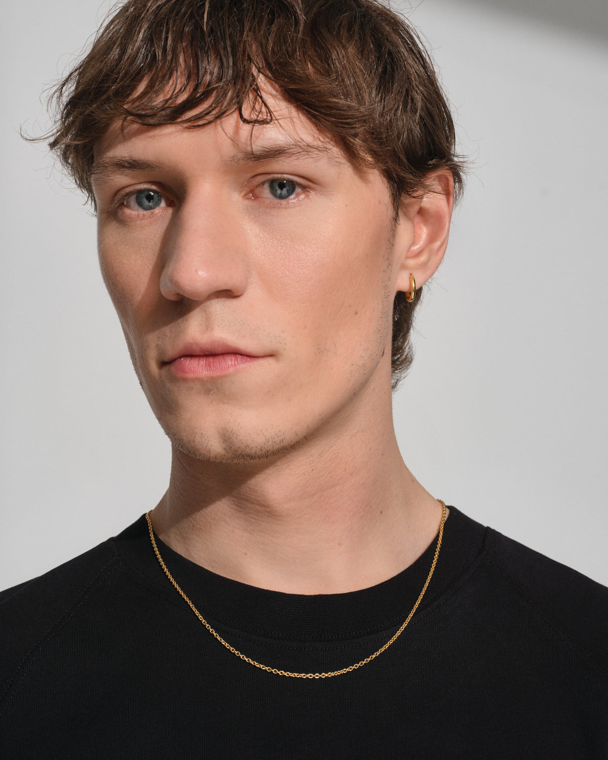 A close-up portrait of a man with light blue eyes and brown hair, wearing a delicate gold chain necklace and a small gold hoop earring, dressed in a black crew-neck shirt against a light gray background.