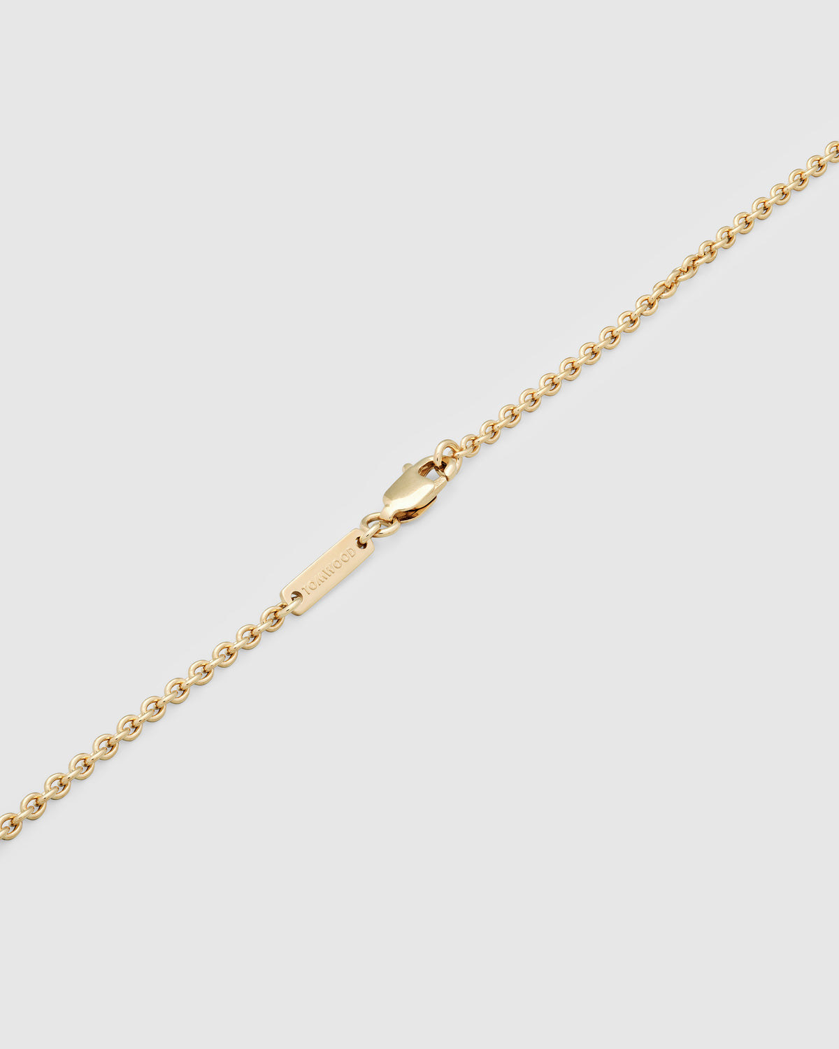A close-up of a gold chain necklace showing the lobster clasp and a small engraved tom wood logo tag on a light gray background.