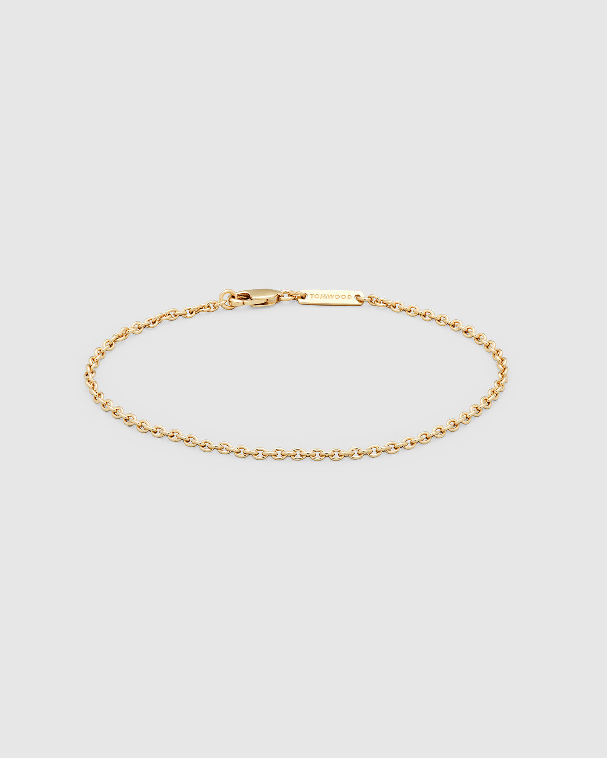 A delicate gold chain bracelet with a lobster clasp and a small rectangular engraved logo tag, displayed on a light gray background.
