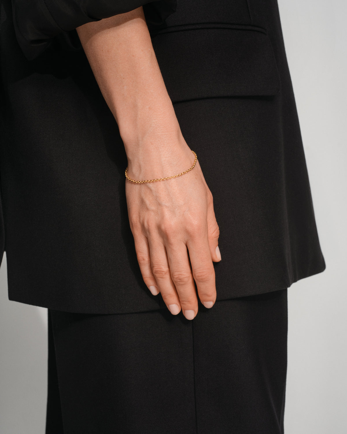 A close-up of a person's hand wearing a delicate gold chain bracelet, styled with a black blazer and black trousers against a light gray background.