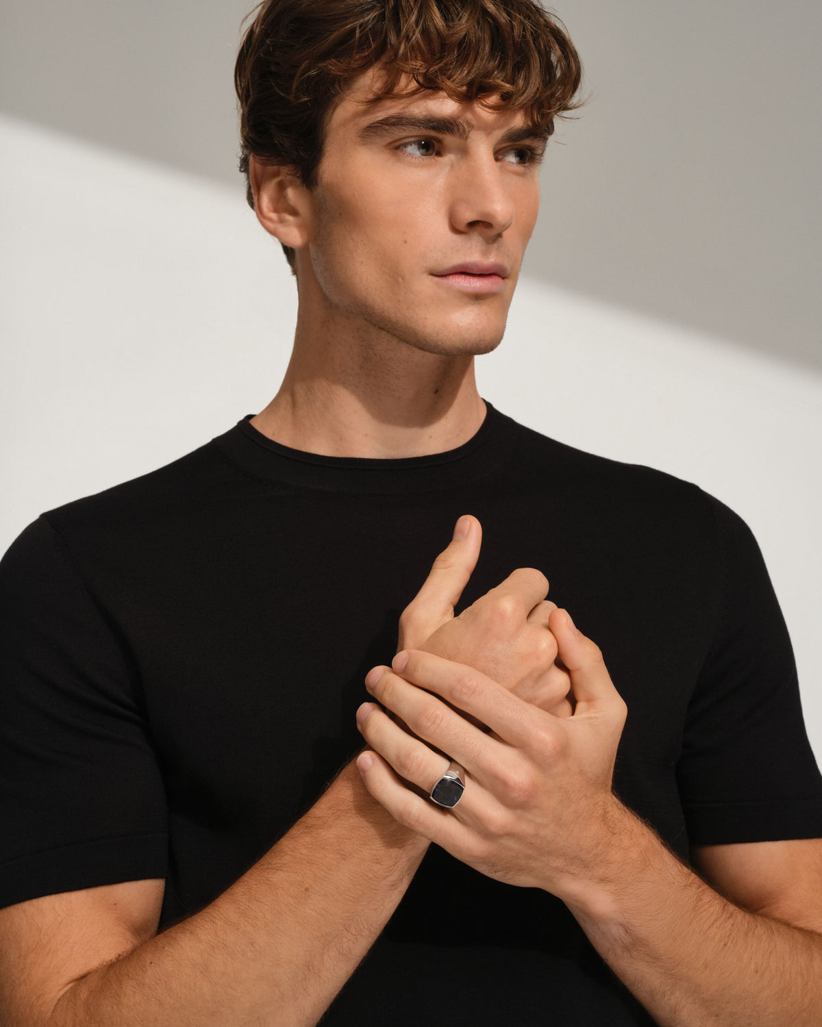 A man wearing a black crew-neck t-shirt and a black ring with a dark pietersite stone, posing with his hands crossed in front of his chest against a light gray background with soft shadows.