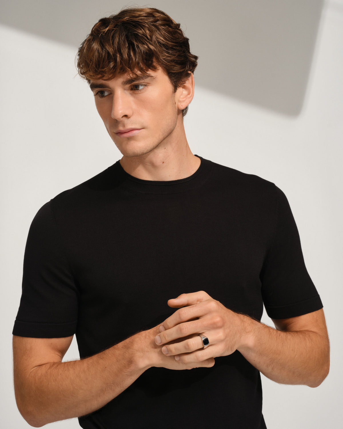A man wearing a black short-sleeve crewneck t-shirt, posing with his hands clasped in front of him against a light gray background with soft shadows.