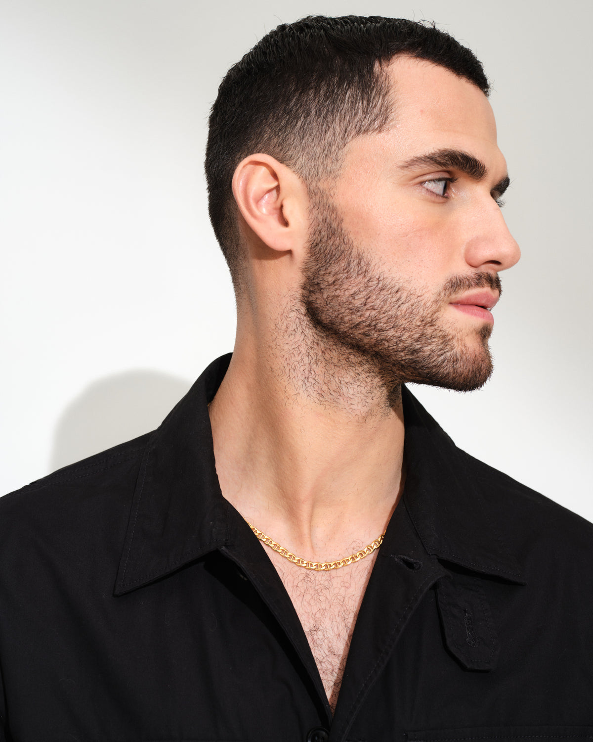 A close-up side profile of a man with short dark hair and a light beard, wearing a black button-up shirt and a gold chain necklace, set against a plain light gray background.